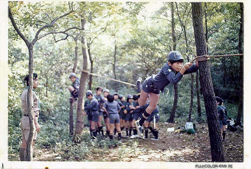 1976年　裏山での訓練　カブ隊による1本ロープ渡りの写真です。 最近はやっていない？ 1本ロープ渡りのコツは片足をロープに引っかけてもう一つの足をブランと下に落としてバランスを取る。できないスカウトもいましたね。 市之瀬パパ隊長が隊長を始めたころ。 ここは通称シカ広場？本当に木々が細い。 この1976年4月に日野2団から分かれた日野5団が生まれています。 スカウトの人数が爆発的に増えた時期です。 そう、正式にシニア隊が同じ4月に発足。